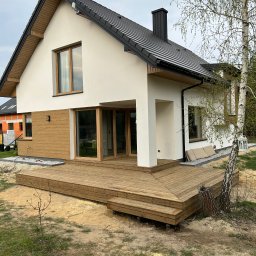 Wooden Harbor Michał Jechna - Nowoczesny dom z białą elewacją i drewnianymi akcentami, z obszernym, wielopoziomowym drewnianym podestem z widocznymi schodkami, otoczony piaszczystym terenem z młodymi drzewami.