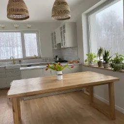 Jasny drewniany stół w minimalistycznej kuchni z widokiem na zimowy krajobraz, rattanowe lampy wiszące i drewniana podłoga.