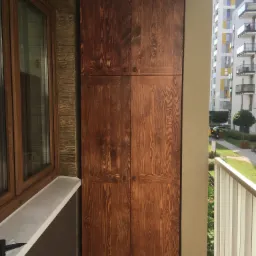Wysoka, drewniana szafa z czterema drzwiczkami na balkonie, widok na osiedle z zielenią.
