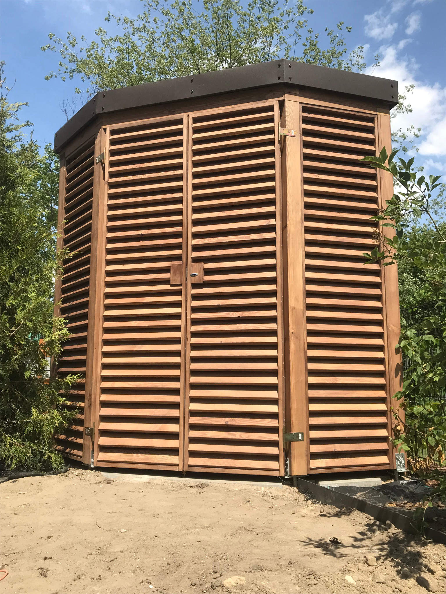 Zamykana, drewniana konstrukcja z żaluzjowymi drzwiami, prawdopodobnie schowek ogrodowy, z brązowym dachem, otoczona zielenią i piaskiem.