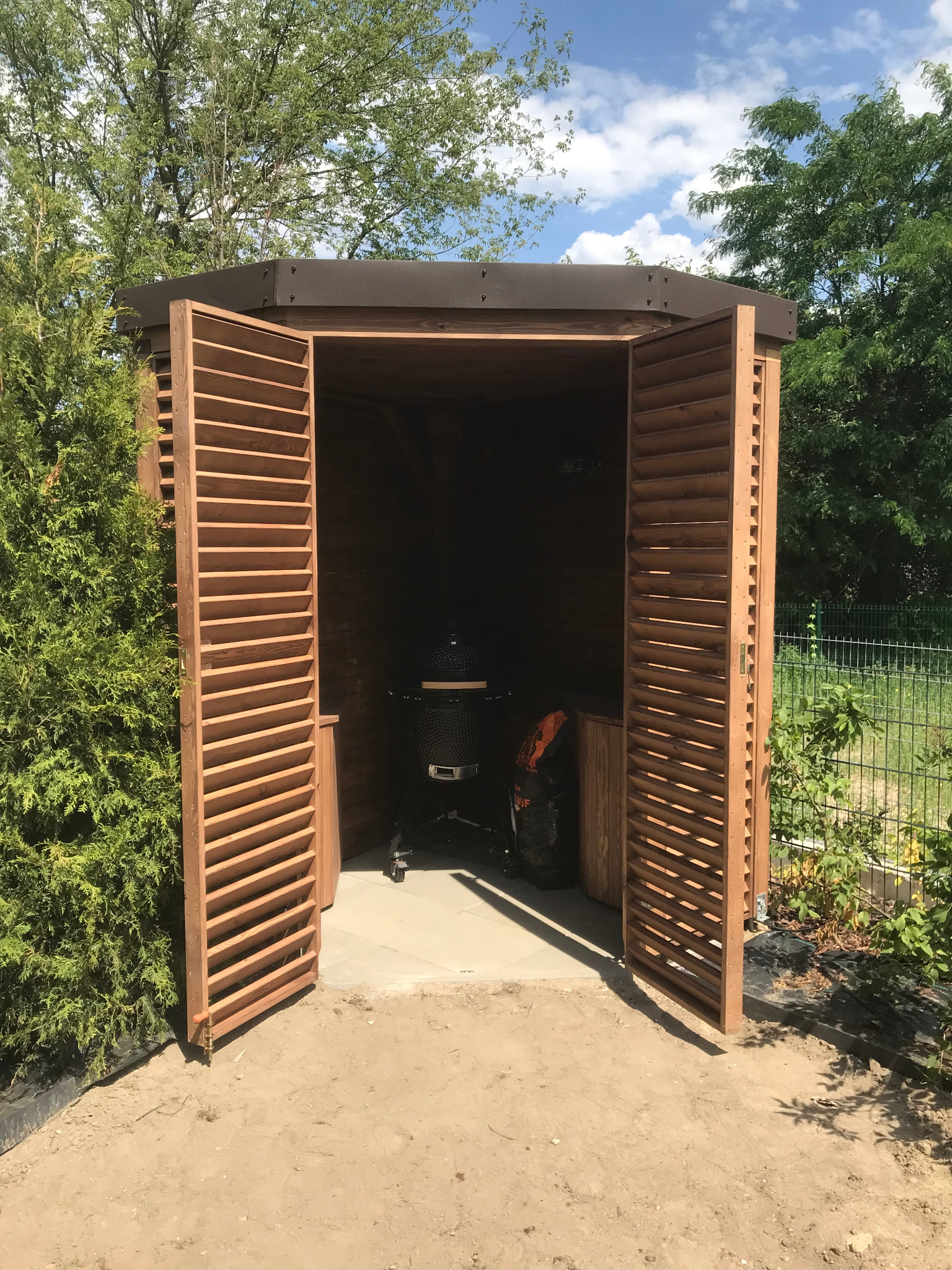 Otwarta drewniana konstrukcja z żaluzjowymi drzwiami, wewnątrz widoczny grill ceramiczny i worek węgla, całość otoczona zielenią.