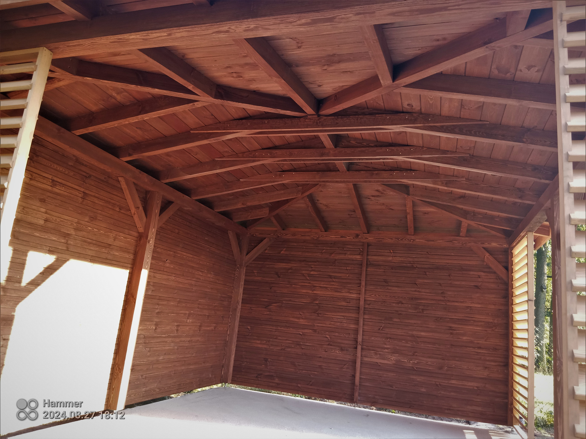 Wnętrze nowej, drewnianej konstrukcji z widoczną więźbą dachową i ścianami obłożonymi boazerią, z elementami ażurowymi po bokach.