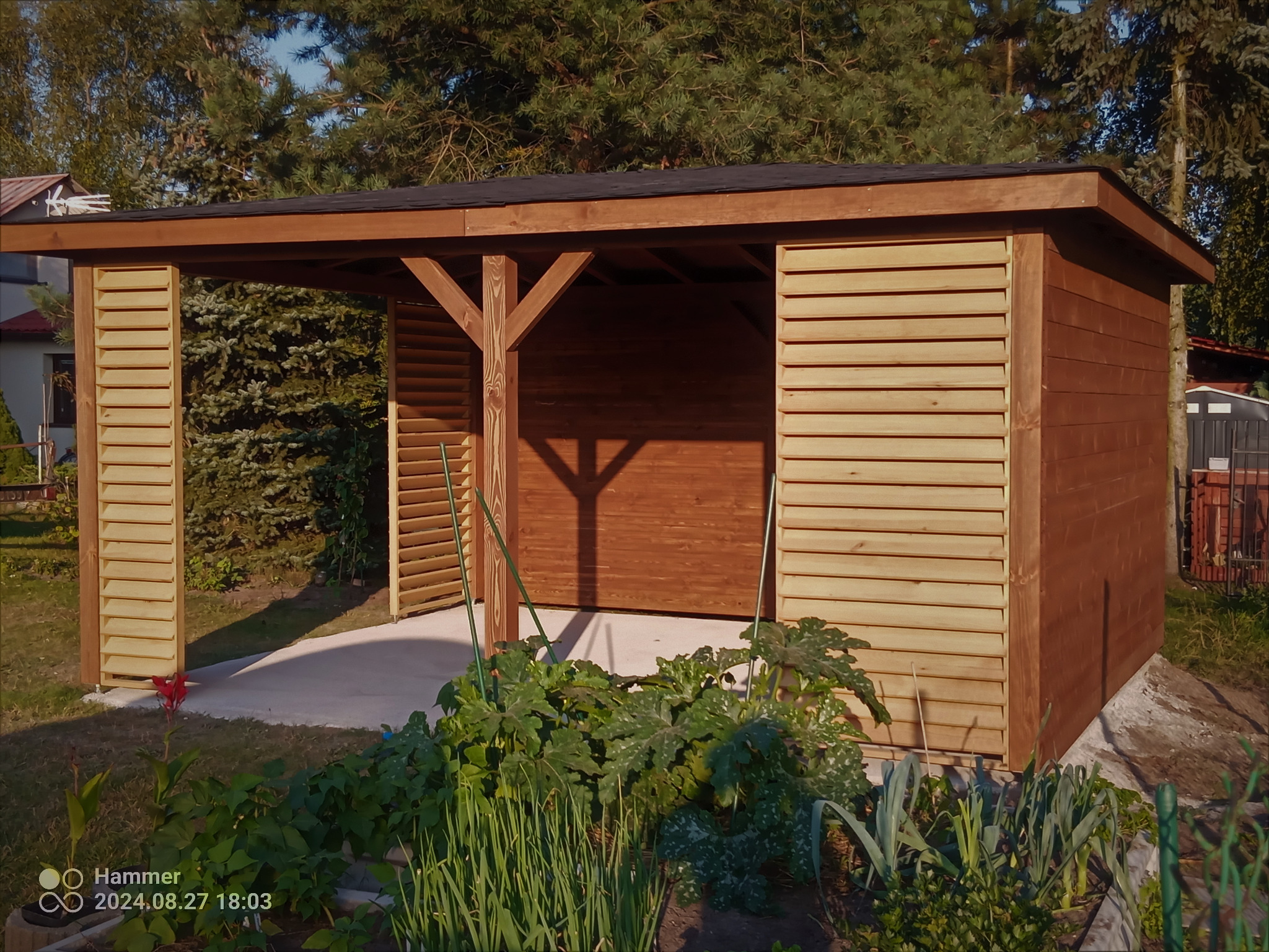 Drewniana konstrukcja z zadaszeniem, prawdopodobnie wiata lub mała altana, z lamelowymi ścianami bocznymi i drewnianymi słupami, otoczona zielenią ogrodową w słoneczny dzień.