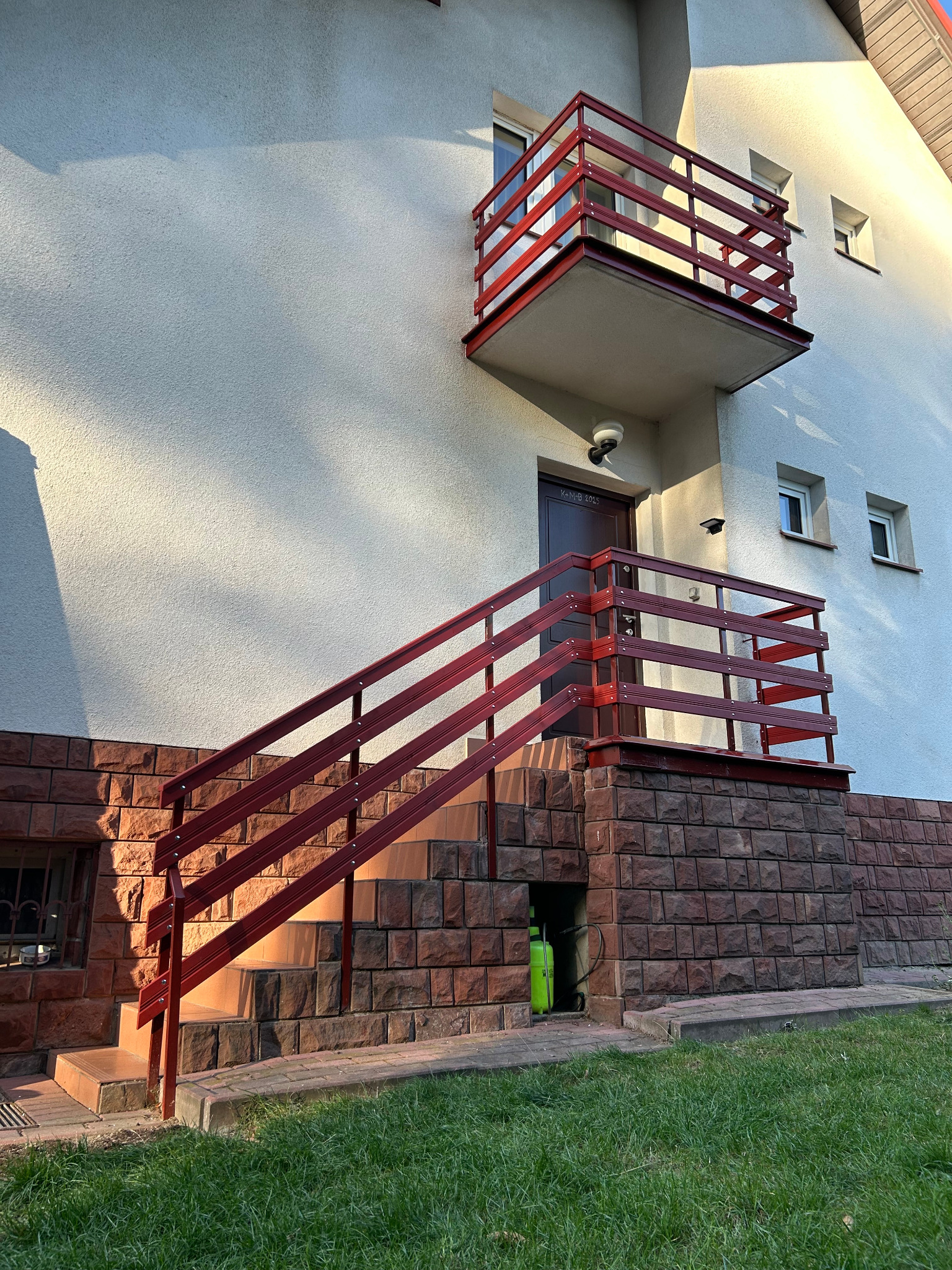 Czerwone balustrady schodów i balkonu przy jasnym budynku z ceglaną podmurówką w słoneczny dzień. Widoczny fragment trawnika.