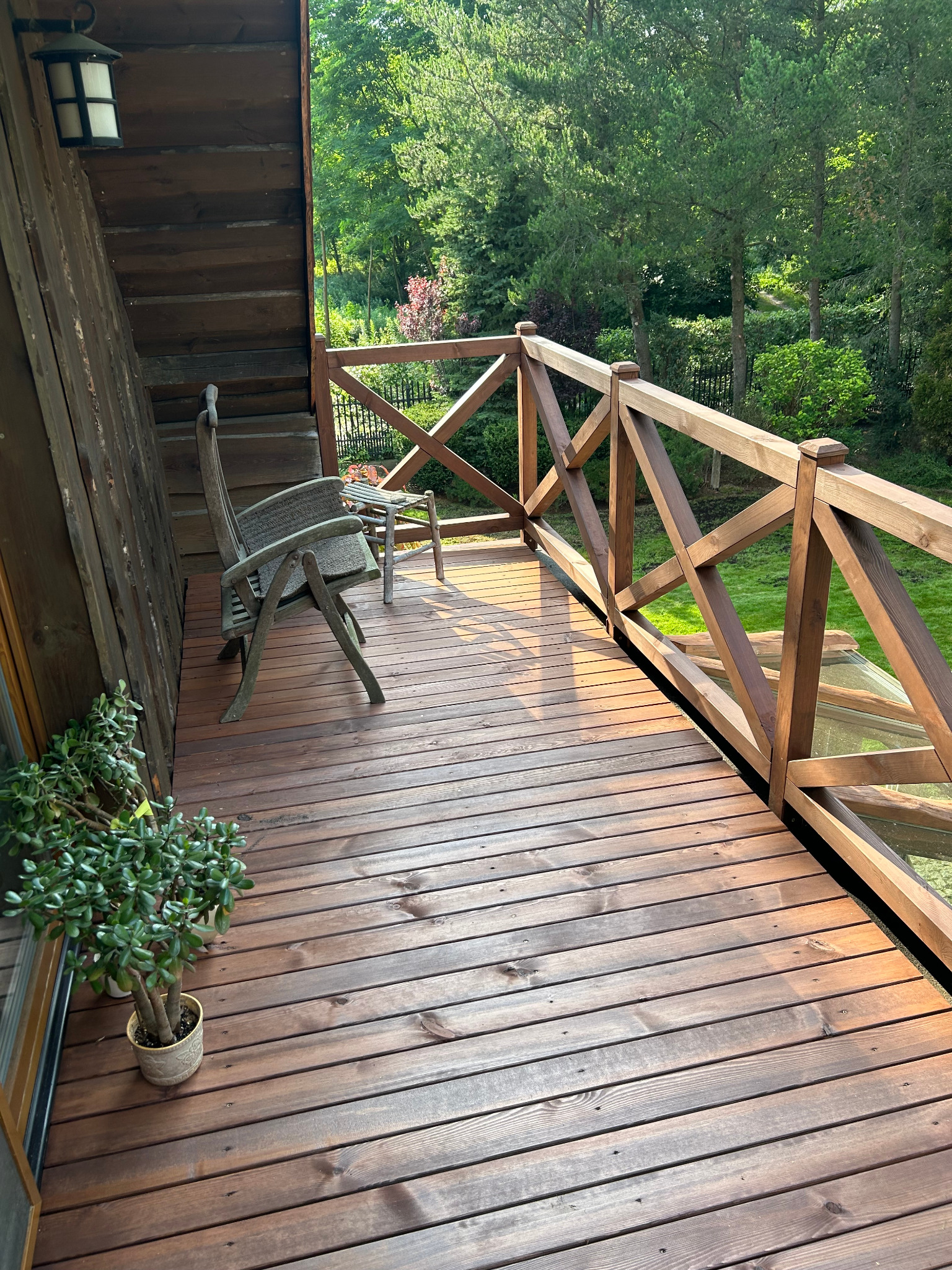 Rustykalny balkon z drewnianą podłogą i balustradą krzyżową, oświetlony ciepłym światłem słonecznym, z krzesłem i widokiem na zieleń.