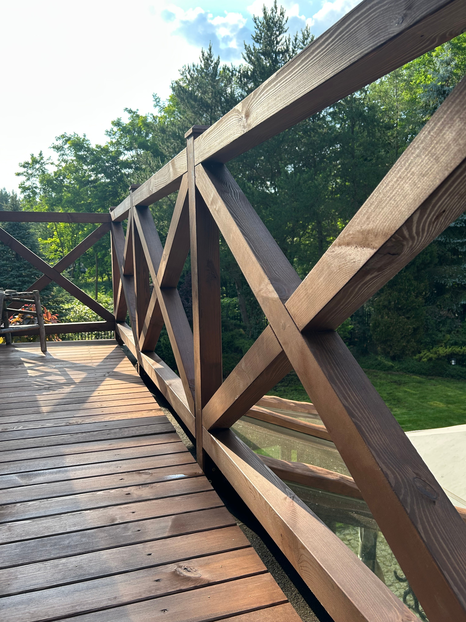 Drewniana balustrada z krzyżującymi się elementami na tarasie, w tle zieleń drzew i krzewów. Widoczne deski podłogowe i fragment mebla ogrodowego.