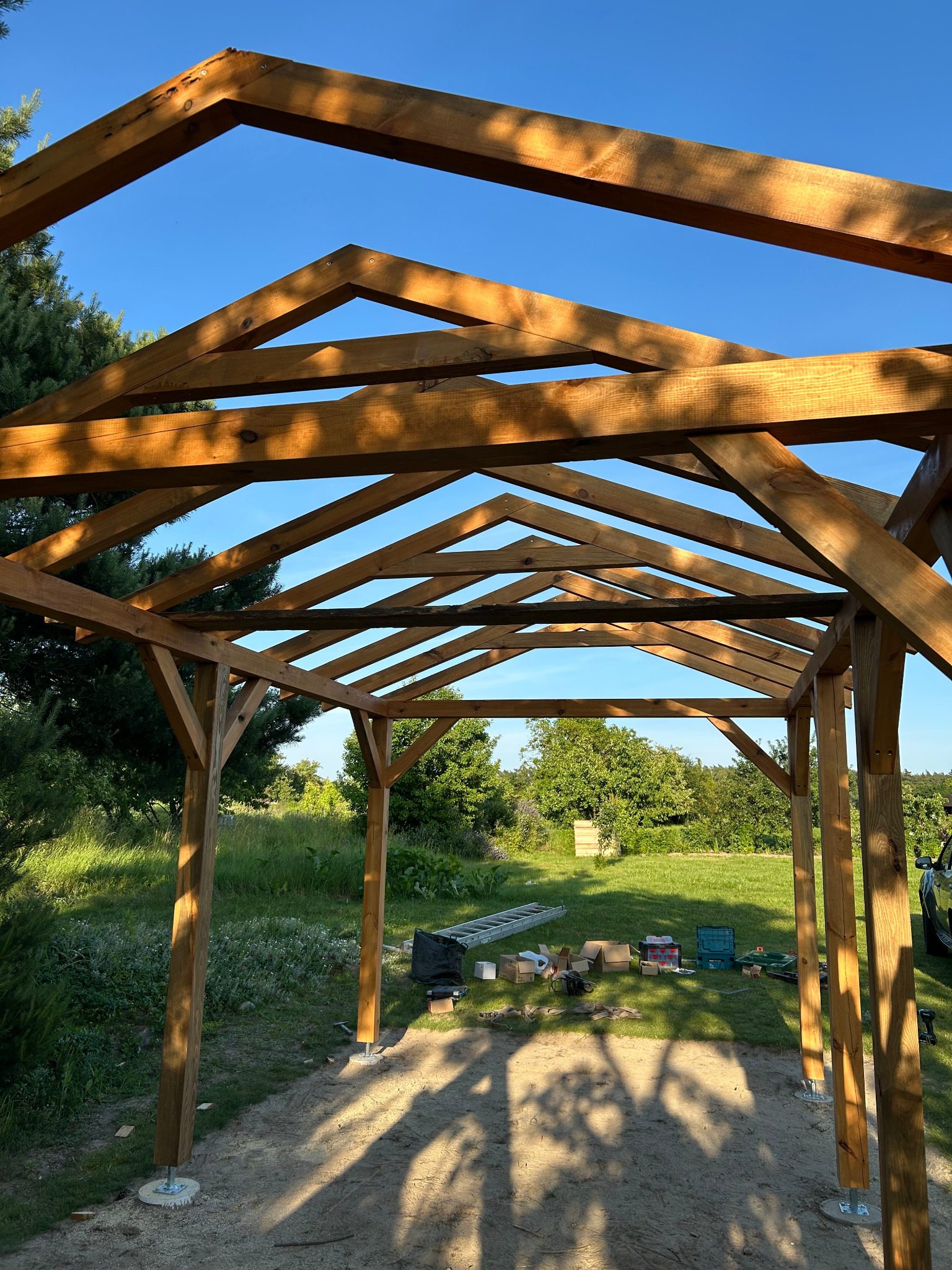 Konstrukcja drewniana altany w trakcie budowy, widoczne słupy i elementy dachu, narzędzia na trawie, słoneczny dzień.