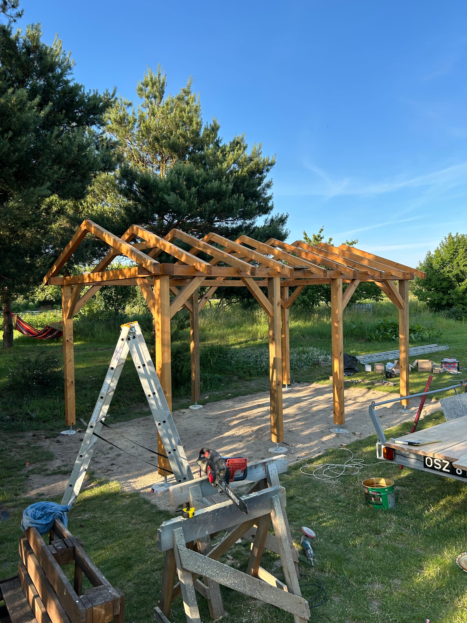 Wznoszenie drewnianej altany: konstrukcja szkieletowa z widocznymi narzędziami na trawniku, drabina oparta o filar, błękitne niebo w tle. Etap budowy.