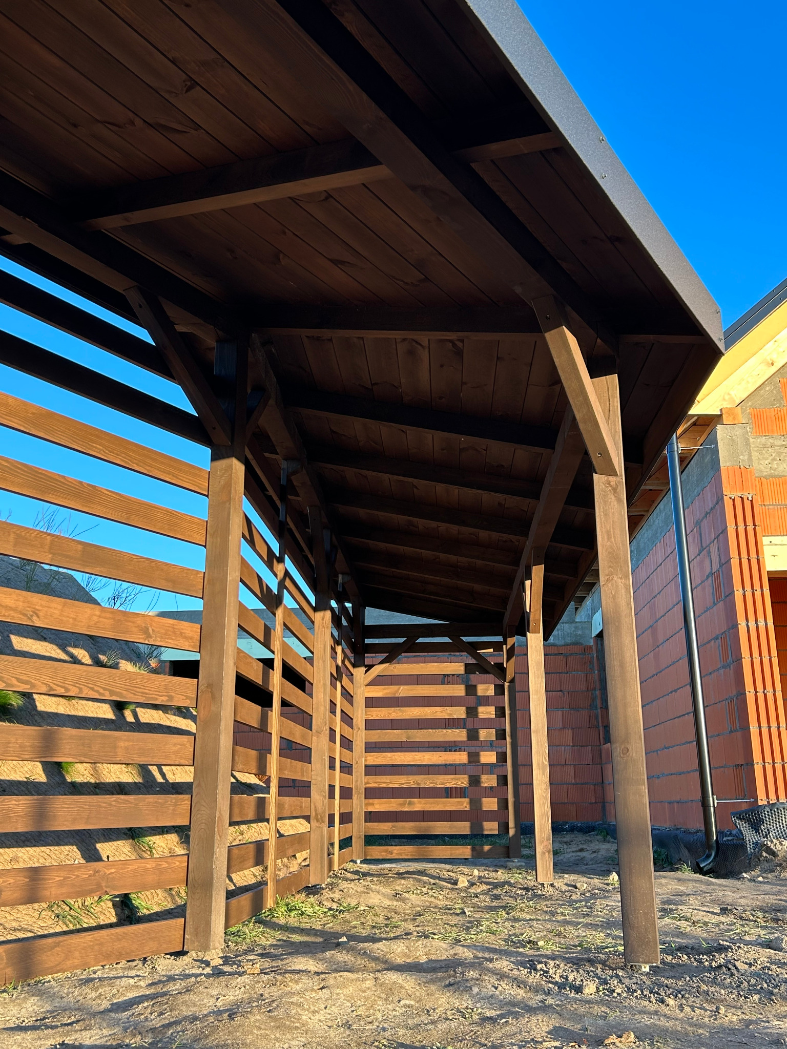 Ciemnobrązowa konstrukcja z drewna, prawdopodobnie wiata, z poziomymi deskami po bokach, na tle błękitnego nieba i budynku w trakcie budowy.