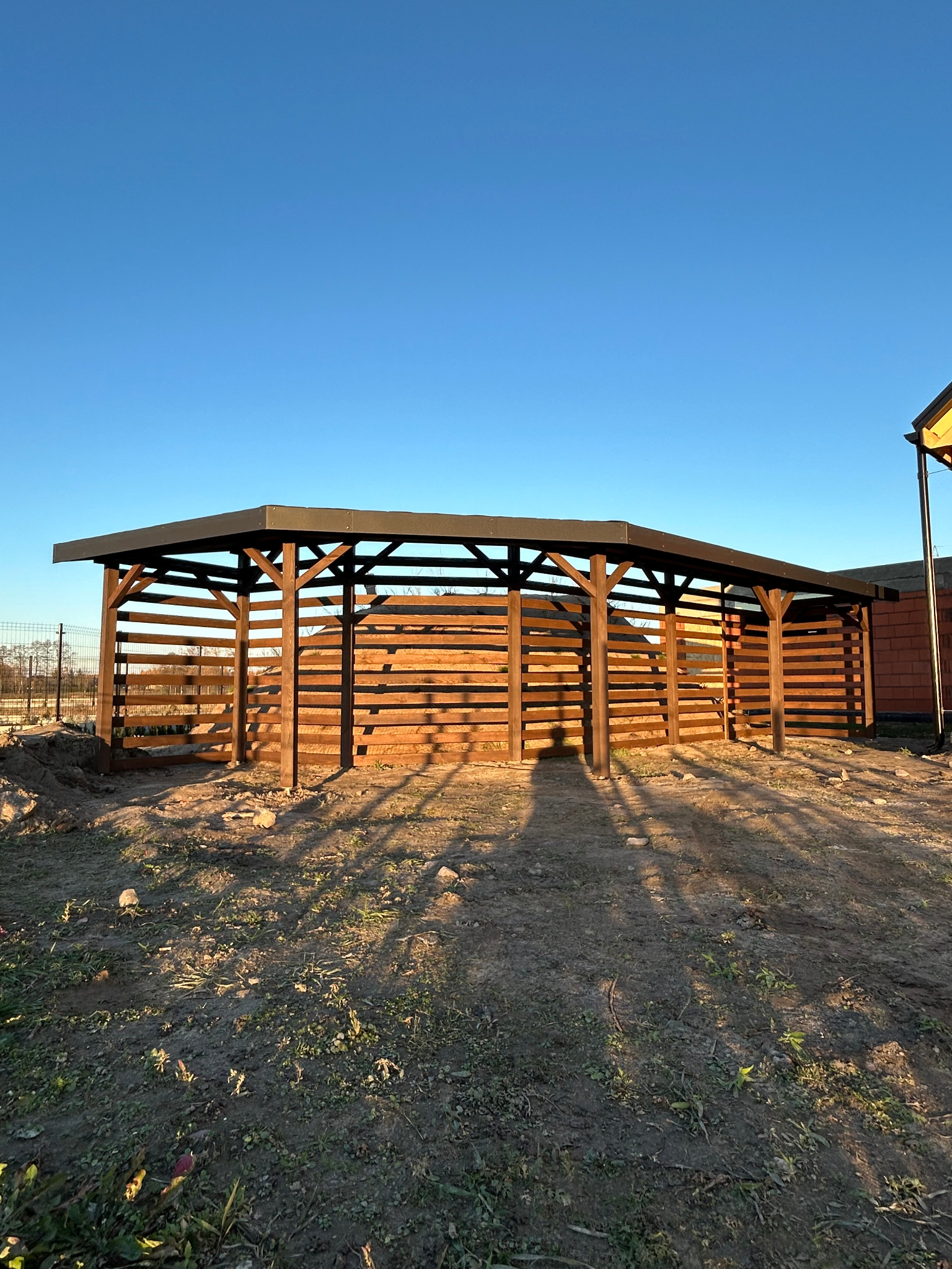 Brązowa konstrukcja z drewna z płaskim dachem na zewnątrz w słoneczny dzień. Widoczne cienie rzucane przez konstrukcję na ziemię. W tle błękitne niebo i fragment budynku.