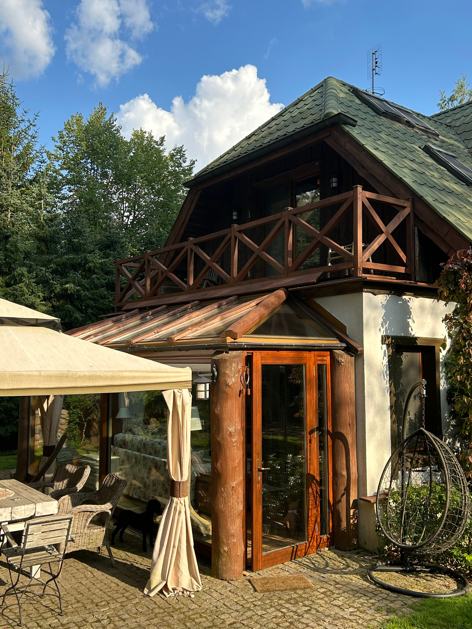 Dom z drewnianą konstrukcją balkonu i przeszklonym tarasem, otoczony zielenią. Widoczny fragment altany i huśtawka ogrodowa. Uwagę przykuwa pies wewnątrz przeszklonej konstrukcji.