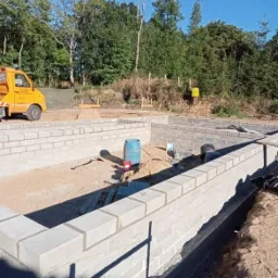 Fundamenty budynku w trakcie budowy, wykonane z szarej kostki betonowej, widoczny żółty samochód dostawczy w tle i drzewa.