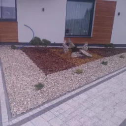 Kwadratowa rabata z kory, kamieni i roślin ozdobnych, obramowana kostką brukową, na tle białej ściany budynku z drewnianą okładziną.