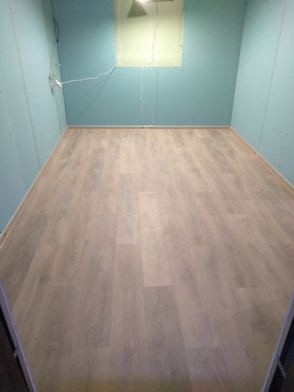 Pokój z wykończonymi ścianami z płyty gipsowo-kartonowej w odcieniach błękitu i nową podłogą z paneli imitujących jasne drewno; widoczne okablowanie elektryczne.