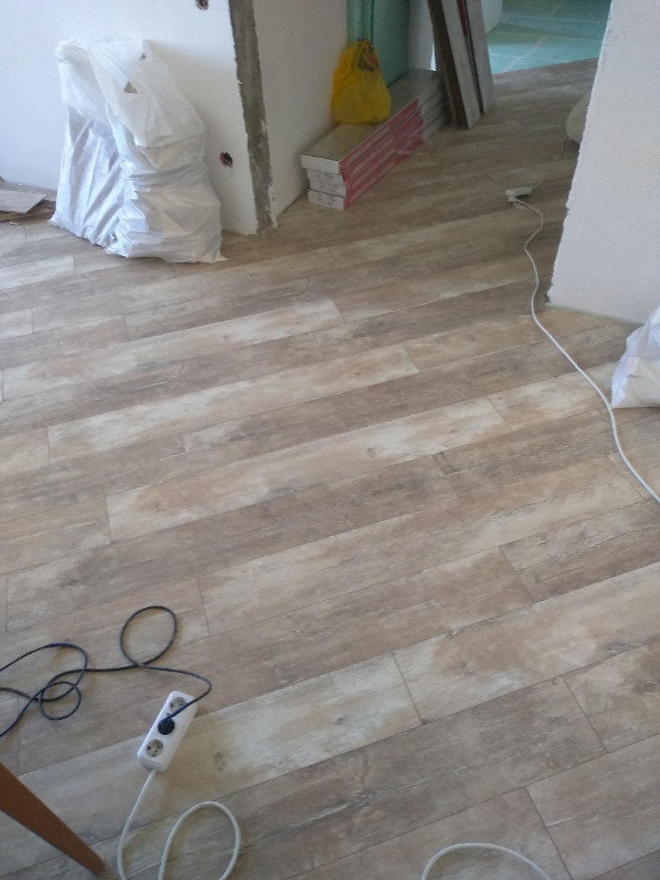 Ułożona podłoga z paneli imitujących drewno, widoczna listwa przypodłogowa, przewody elektryczne i materiały budowlane w workach i kartonach podczas prac wykończeniowych.