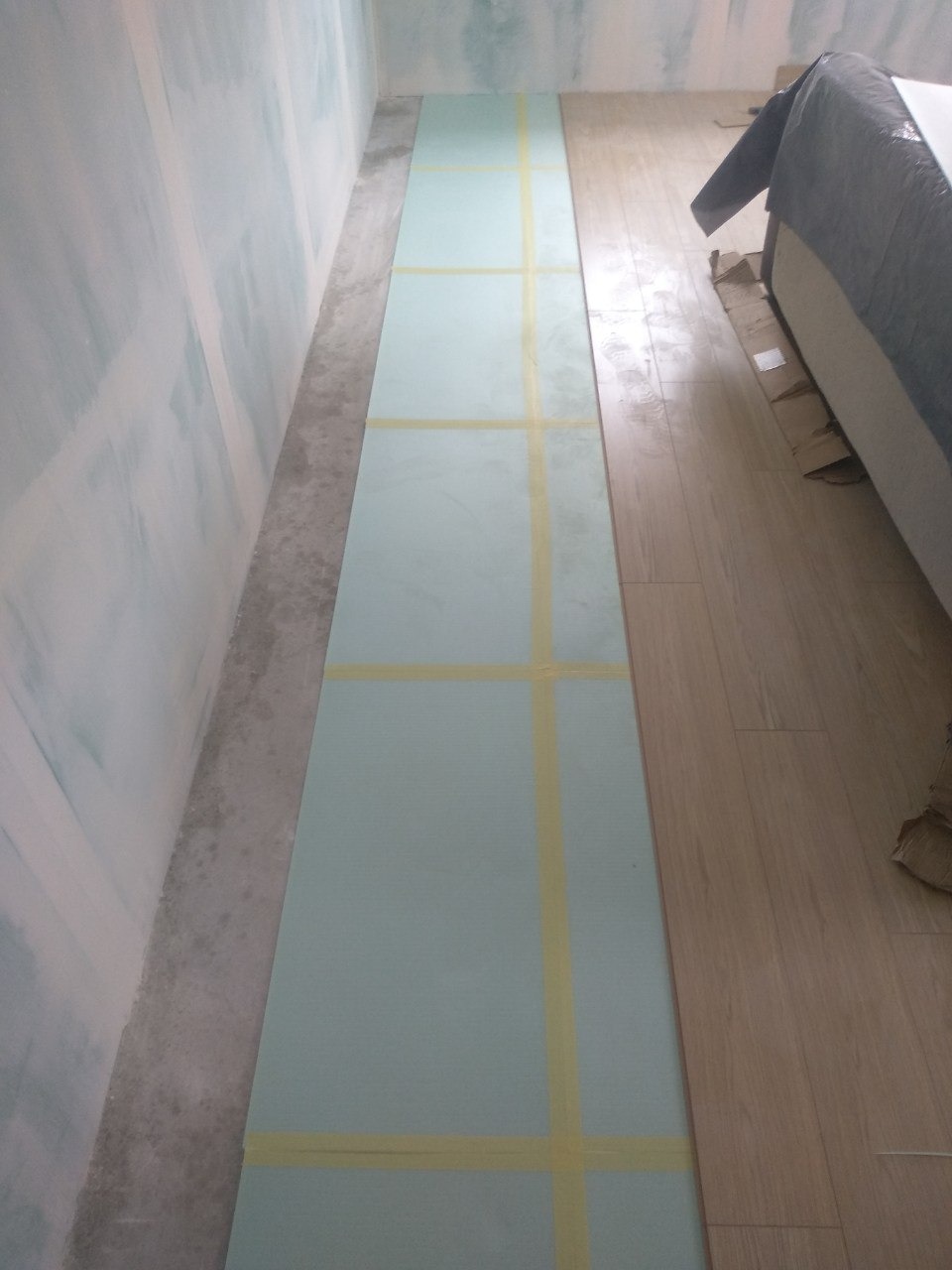 Przygotowanie podłoża pod parkiet: płyta izolacyjna z taśmą klejącą na podłodze, widoczna ściana po obróbce gipsowej i fragment ułożonej podłogi drewnianej.