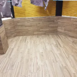 Wykończone wnętrze z podłogą i ścianami wyłożonymi płytkami imitującymi drewno, widoczne zwisające kable i fragment zwiniętej rolety okiennej.