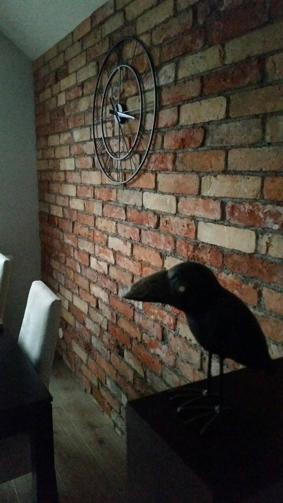 Dekoracyjna ściana z czerwonej cegły z metalowym zegarem w stylu industrialnym i figurką kruka na ciemnej komodzie.