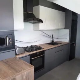 Nowoczesna kuchnia w odcieniach szarości i bieli, z czarnym okapem i sprzętem AGD, drewnopodobny blat, minimalistyczny design.