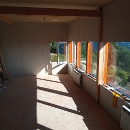 Brak - Wnętrze w trakcie wykańczania: płyty karton-gips na ścianach, drewniane ramy okienne, grzejniki, podłoga z płyt OSB, oświetlone promieniami słońca. Widok na zieleń za oknem.