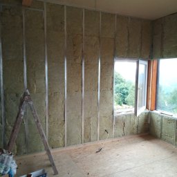 Brak - Wnętrze w trakcie izolacji: wełna mineralna między metalowymi profilami na ścianach, drewniana podłoga, otwarte okno z widokiem na zieleń. Widoczna drabina i karton.