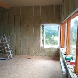 Brak - Ocieplenie ścian wełną mineralną z metalowym stelażem, widoczna drabina i okno z widokiem na zieleń. Podłoga z płyt OSB, grzejnik przy oknie.