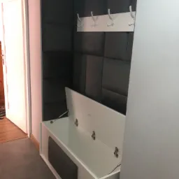 Wnęka z siedziskiem i wieszakami w przedpokoju, szara tapicerowana ściana w tle, otwarte siedzisko z szarą wstawką.