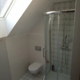 Wykończona łazienka na poddaszu ze skośnym oknem dachowym, kabiną prysznicową z wzorzystą ścianą i podwieszaną toaletą.