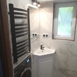 Nowoczesna łazienka z umywalką narożną na szafce, grzejnikiem drabinkowym obok pralki i marmurowymi płytkami na ścianach.