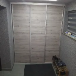 Trzydrzwiowa szafa wnękowa z przesuwnymi drzwiami o fakturze jasnego drewna, oświetlenie LED w suficie, dywanik i półka na buty przy ścianie o szarej, ziarnistej fakturze.