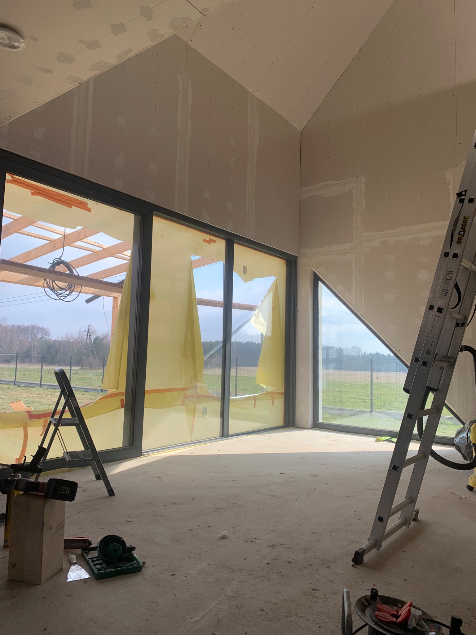 Wnętrze w trakcie wykańczania: ściany z płyt karton-gips, duże okna z widokiem na zewnątrz, drabiny i narzędzia budowlane na podłodze. Charakterystyczne skośne ściany.