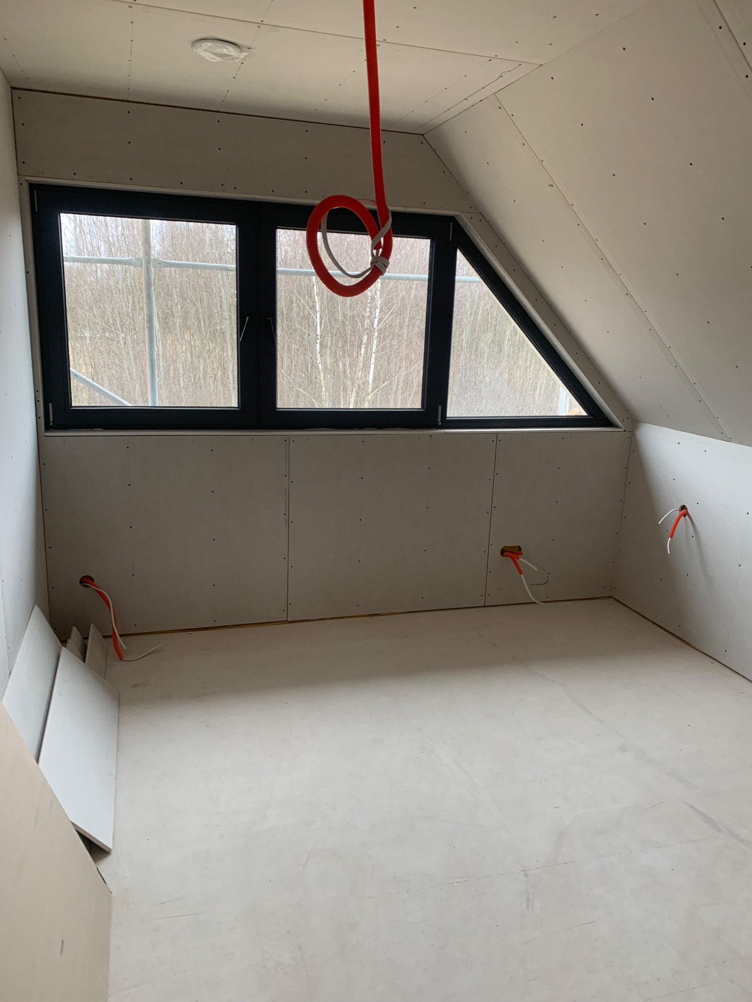 Poddasze z oknem i zamontowaną płytą karton-gips, widoczne kable elektryczne w ścianach i rura instalacyjna pod sufitem. Surowy stan wykończenia wnętrza.