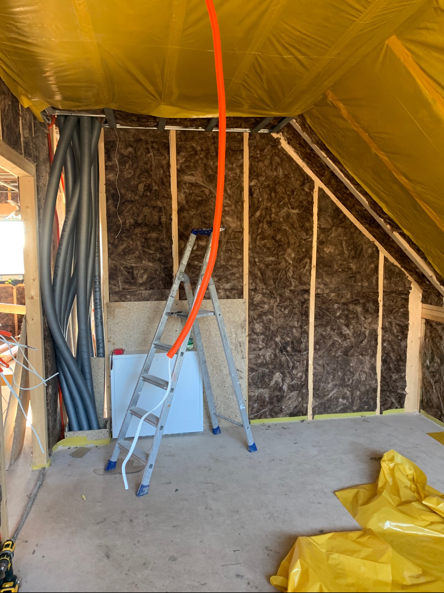 Poddasze w trakcie izolacji: widoczne ocieplenie ścian wełną mineralną, rury instalacyjne, drabina i folia ochronna. Prace wykończeniowe w surowym stanie.