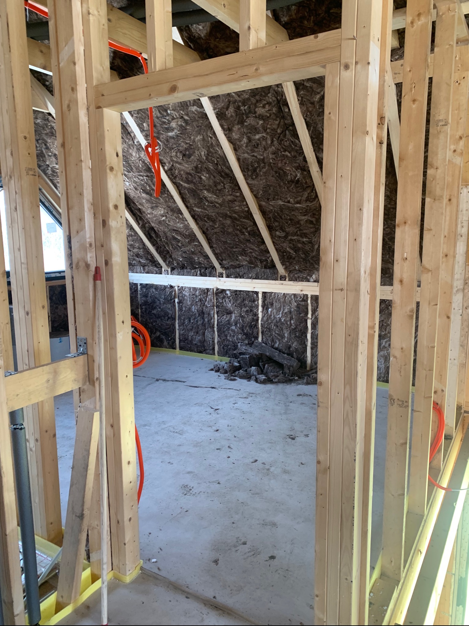 Konstrukcja szkieletowa poddasza z izolacją, widoczne drewniane belki, instalacja elektryczna w peszlach i fragment okna. Prace wykończeniowe w surowym stanie.