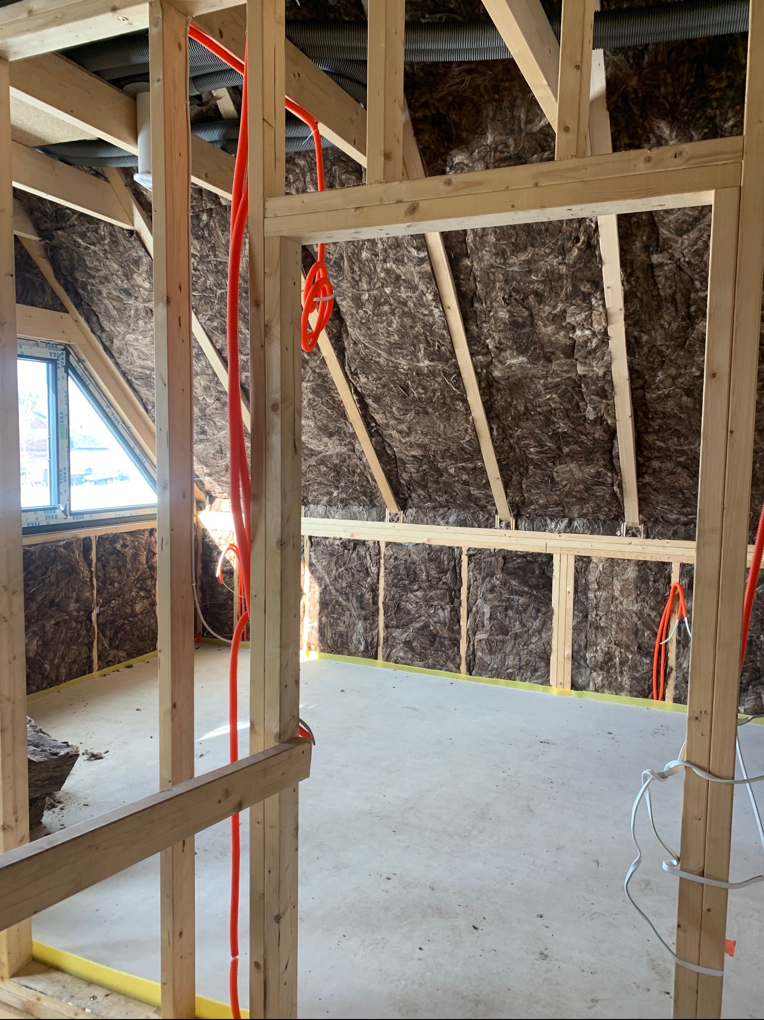 Poddasze w trakcie adaptacji: drewniana konstrukcja, izolacja z wełny mineralnej, instalacja elektryczna w peszlu. Widok na okno dachowe i fragment budynku na zewnątrz.