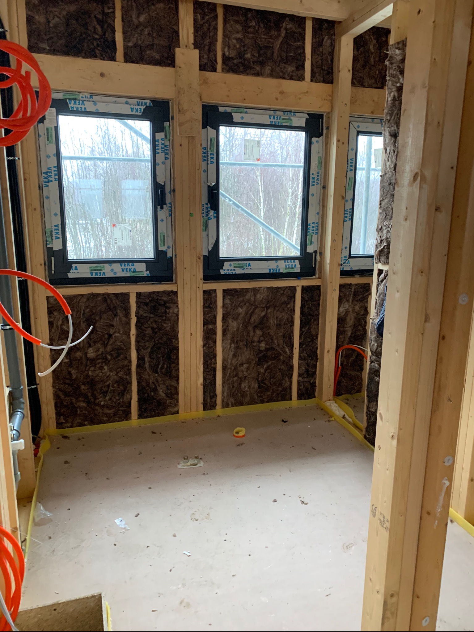 Surowy stan łazienki w trakcie remontu: widoczne okna Veka, izolacja ścian wełną mineralną, drewniana konstrukcja szkieletowa i instalacje hydrauliczne w rurach.