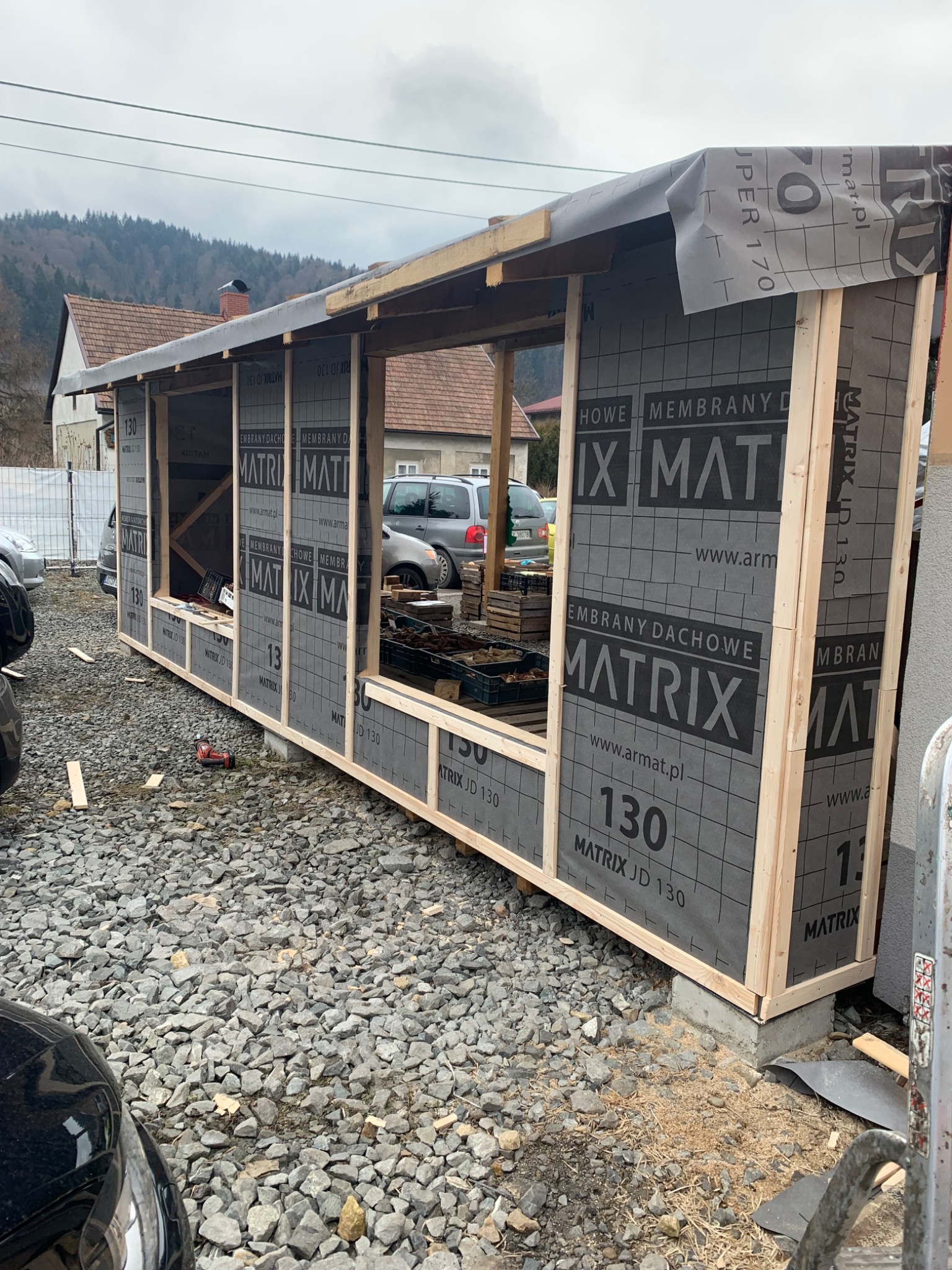 Konstrukcja szkieletowa z membraną dachową Matrix JD 130, widok z zewnątrz na placu budowy. Elementy drewniane, szara membrana, podłoże z kamieni.