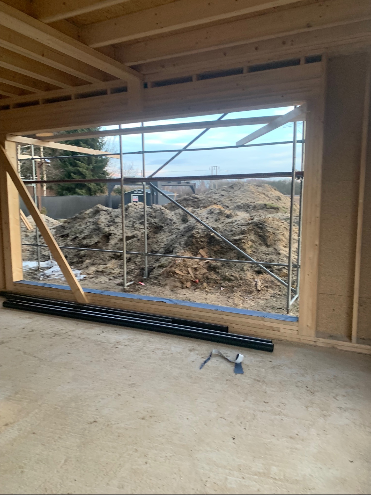 Surowy stan: drewniana konstrukcja okienna w trakcie budowy, widok na zewnątrz z rusztowaniami i stertą ziemi. Rury i narzędzia na podłodze.