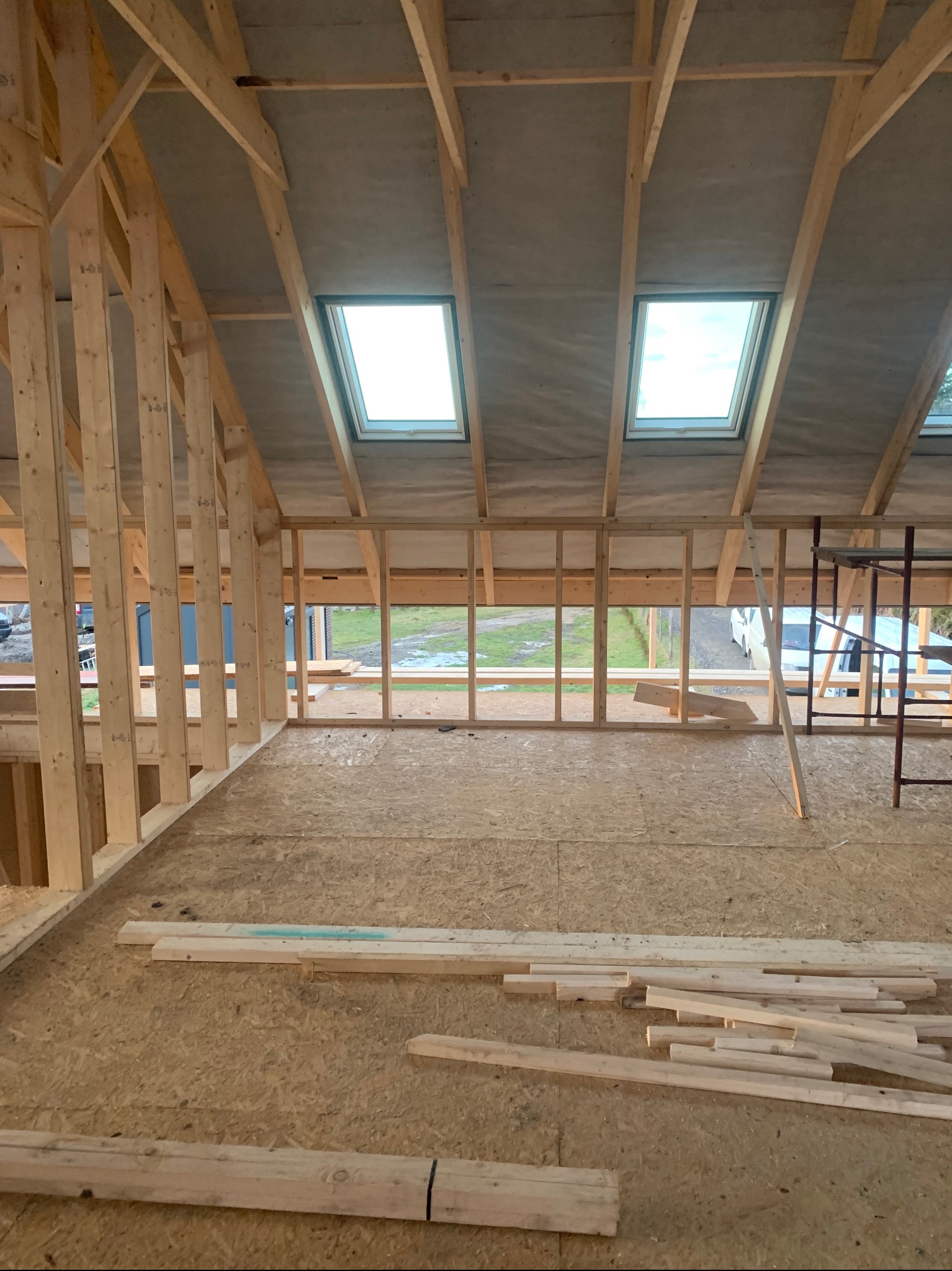Surowe wnętrze poddasza z drewnianą konstrukcją szkieletową, oknami dachowymi i płytami OSB na podłodze. Widoczne elementy rusztowania i przygotowane deski.