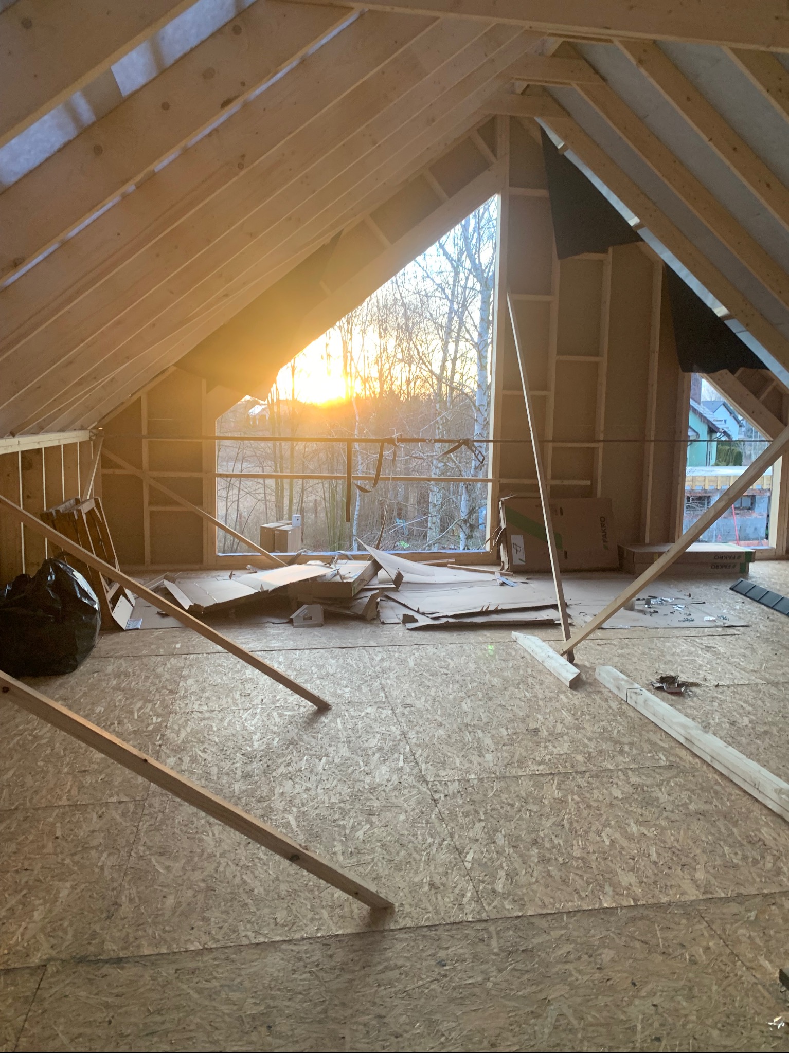 Surowe poddasze z drewnianą konstrukcją dachu i ścian, widok na zachód słońca przez okno. Na podłodze płyty OSB, resztki materiałów budowlanych i narzędzia.
