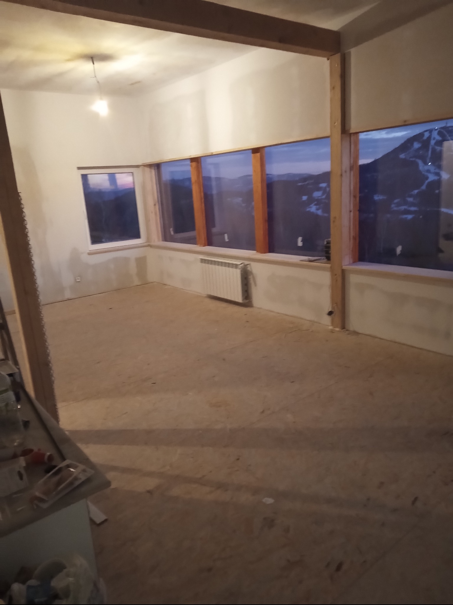 Surowy stan wnętrza z dużymi oknami z widokiem na góry, grzejnik pod oknem, drewniane elementy konstrukcyjne, przygotowany do dalszych prac wykończeniowych.