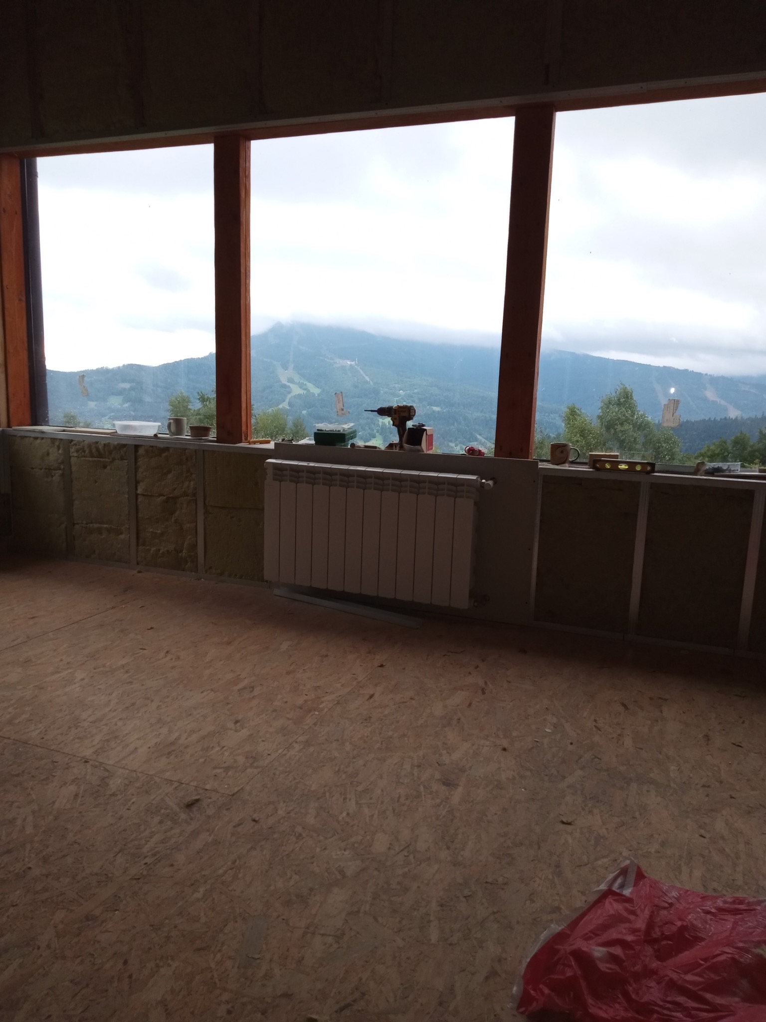 Wnętrze w trakcie prac wykończeniowych: ocieplone ściany z widoczną izolacją, grzejnik, narzędzia na parapecie z malowniczym widokiem na góry w tle.