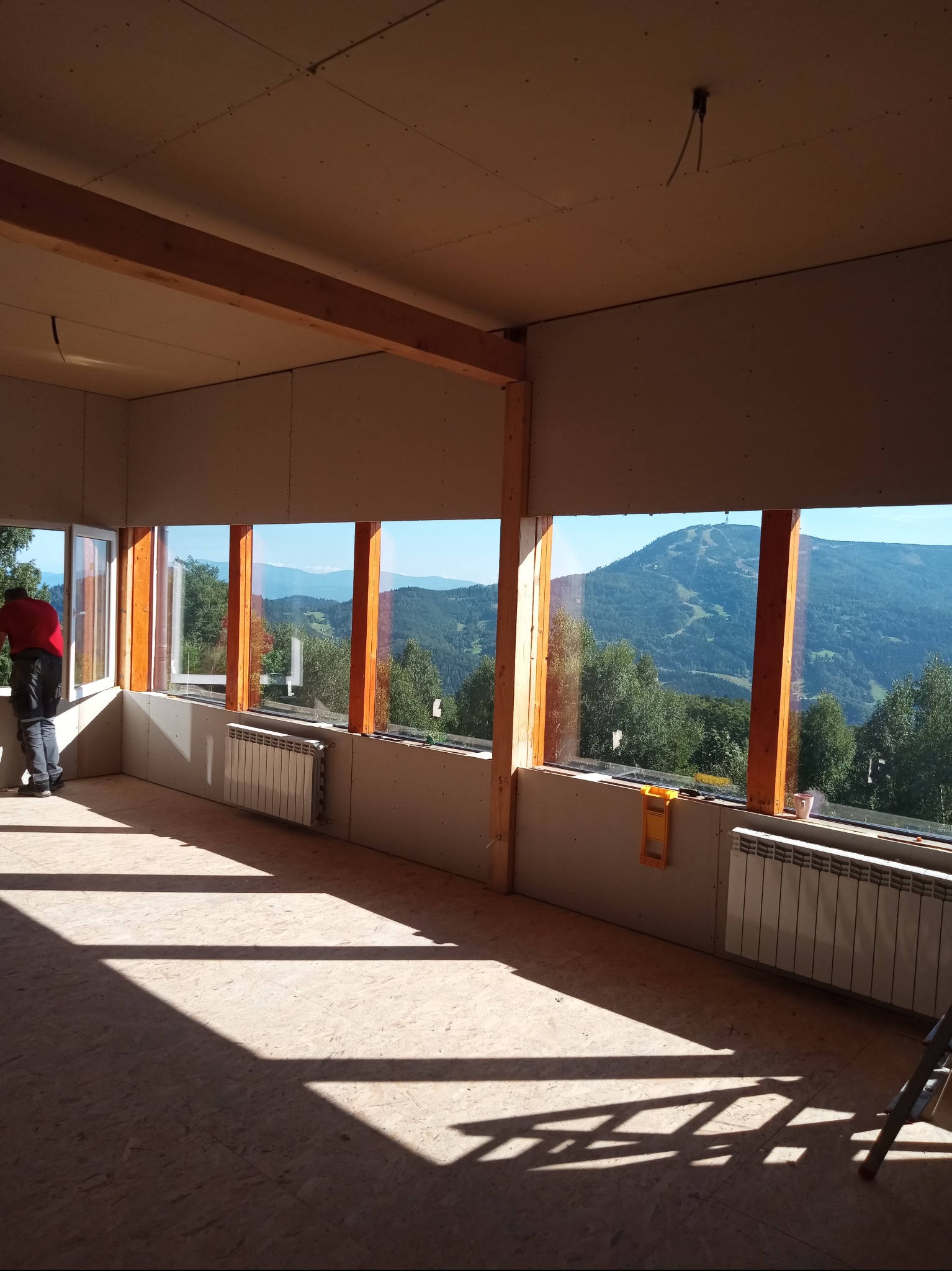 Wnętrze w trakcie wykańczania: płyty karton-gips na ścianach, drewniane ramy okienne z widokiem na góry, grzejnik. Osoba w czerwonej koszulce przy oknie.