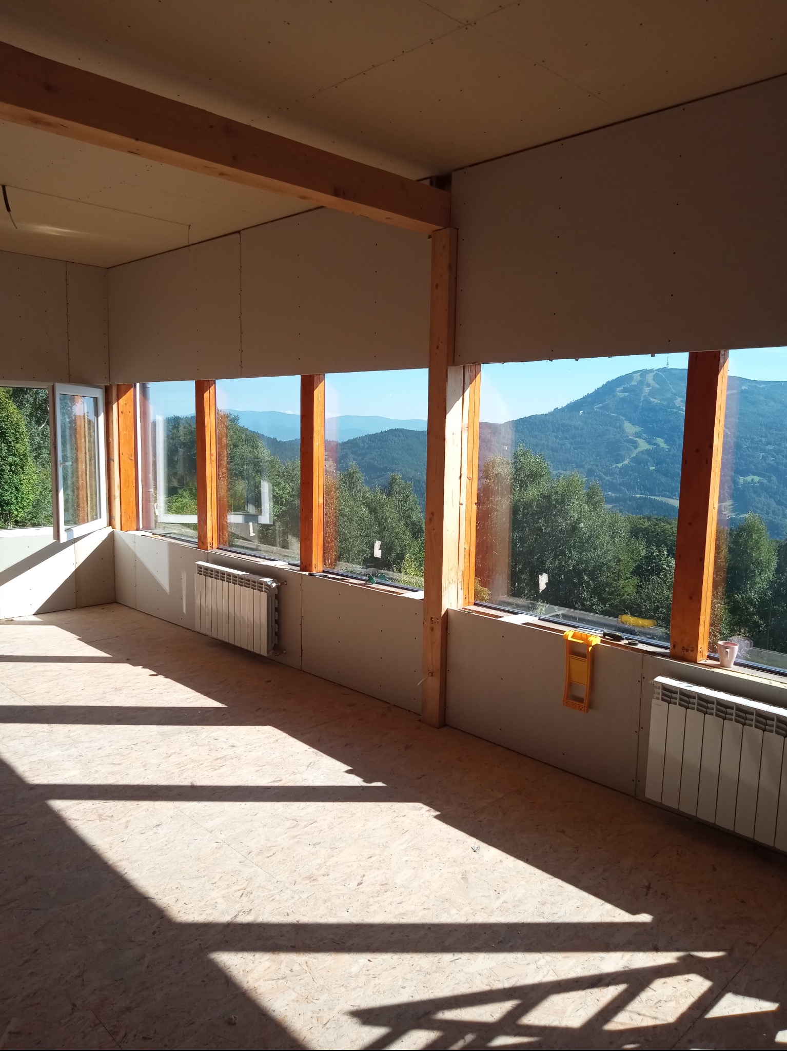 Wnętrze w trakcie wykańczania: płyty karton-gips na ścianach, drewniane ramy okienne z widokiem na góry, grzejniki i narzędzia. Promienie słoneczne wpadają przez okna.