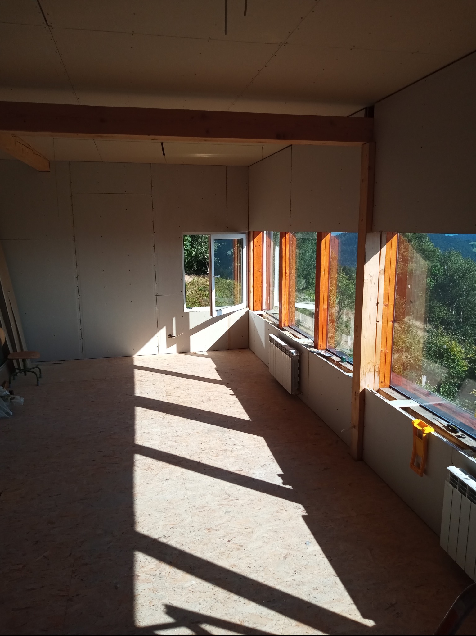 Wnętrze w trakcie wykańczania: płyty karton-gips na ścianach, drewniane ramy okienne, grzejniki, podłoga z płyt OSB, oświetlone promieniami słońca. Widok na zieleń za oknem.