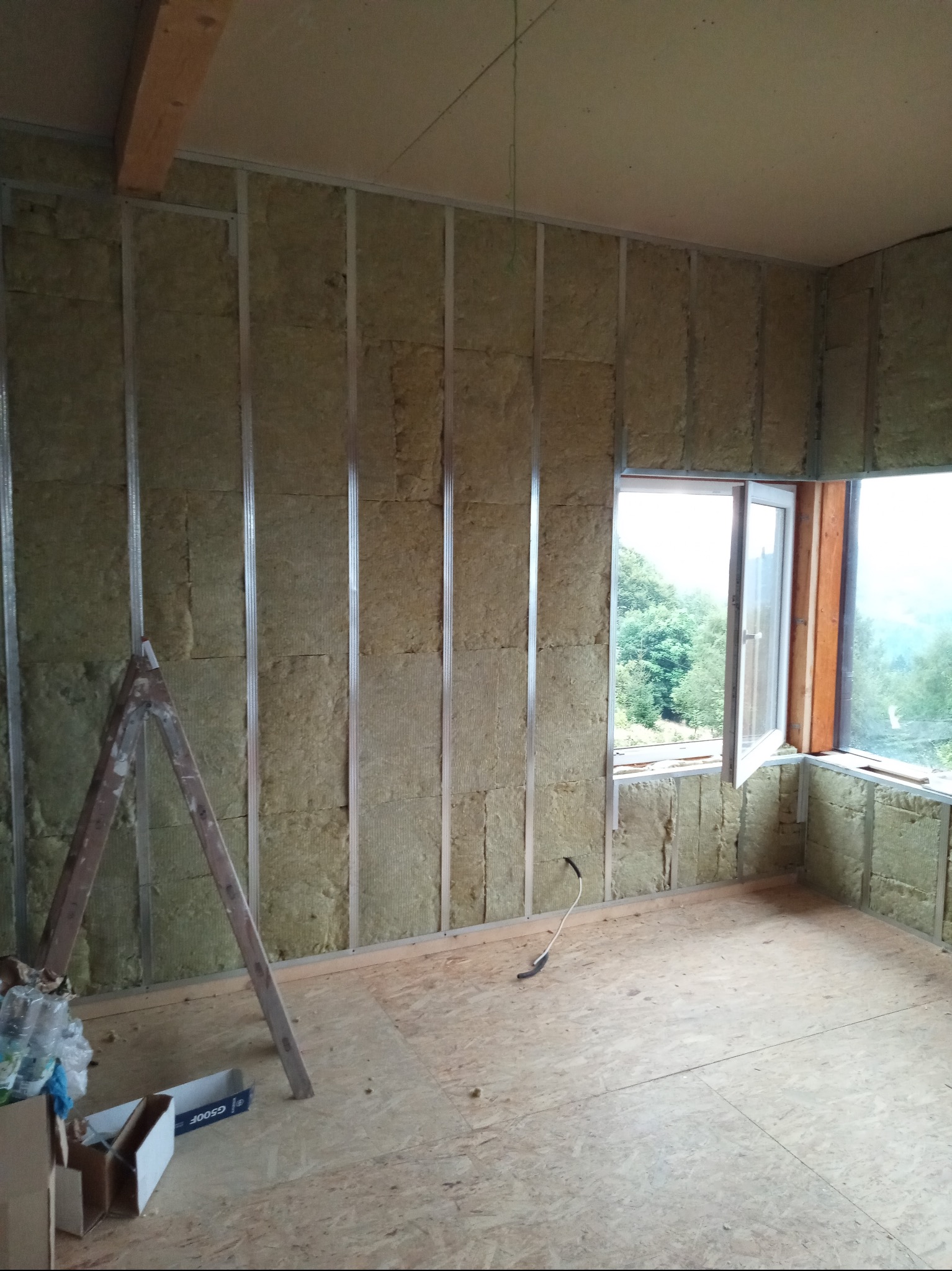 Wnętrze w trakcie izolacji: wełna mineralna między metalowymi profilami na ścianach, drewniana podłoga, otwarte okno z widokiem na zieleń. Widoczna drabina i karton.