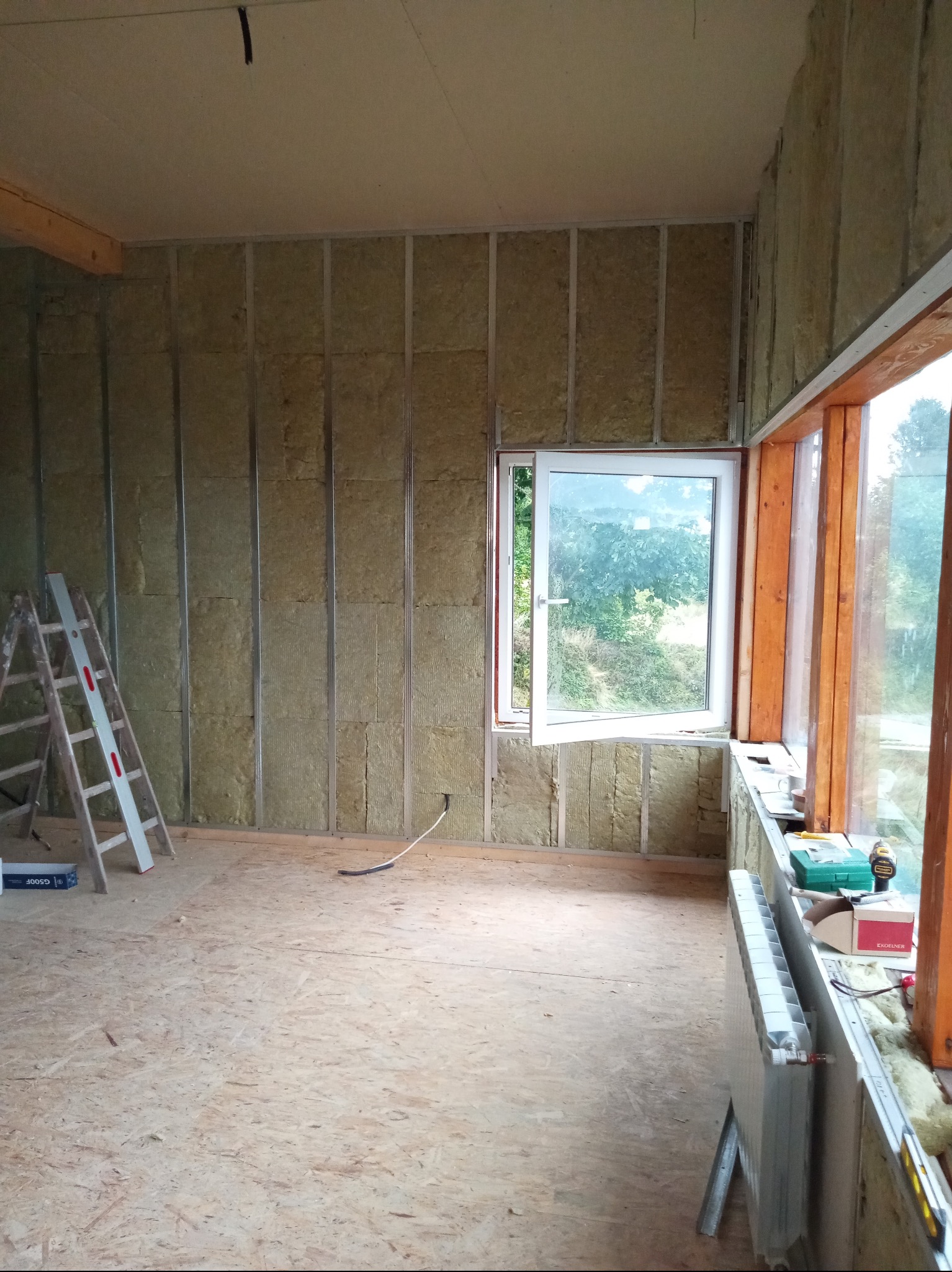 Ocieplenie ścian wełną mineralną z metalowym stelażem, widoczna drabina i okno z widokiem na zieleń. Podłoga z płyt OSB, grzejnik przy oknie.