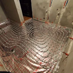 Białe rury ogrzewania podłogowego ułożone spiralnie na folii izolacyjnej z czerwoną siatką w surowym pomieszczeniu łazienki przed wylaniem posadzki.