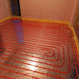 Czerwone rurki ogrzewania podłogowego ułożone na srebrnej folii izolacyjnej w pomieszczeniu z przygotowanymi ścianami.