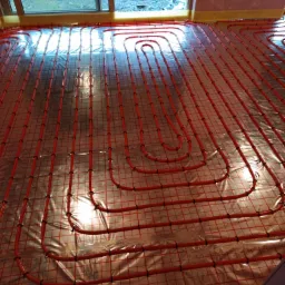 Czerwone rurki ogrzewania podłogowego ułożone na folii z siatką, widoczne okno i fragment ściany